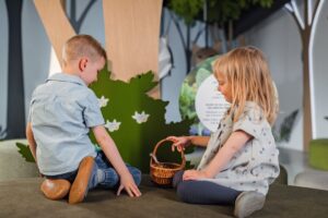 Kleuters spelen rondom een vertel- of thematafel met poppenspel en themamaterialen, begeleid door een leerkracht.