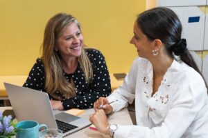 Onderwijsprofessional in gesprek met collega over begeleiding van een leerling, met laptop en notities op tafel.